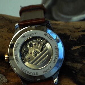 May include: The back of a silver watch with a brown leather band. The watch face is clear and shows the inner workings of the watch. The watch is water resistant to 3 ATM and has a stainless steel back.