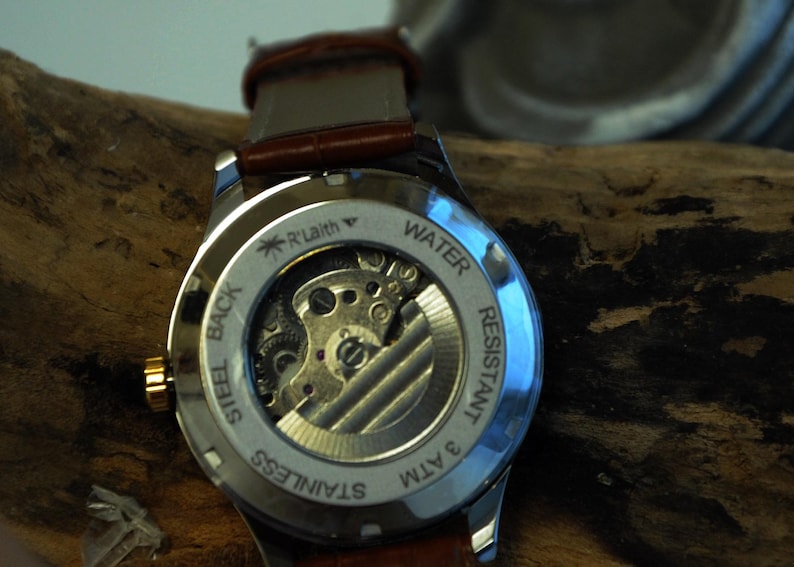 May include: The back of a silver watch with a brown leather band. The watch face is clear and shows the inner workings of the watch. The watch is water resistant to 3 ATM and has a stainless steel back.