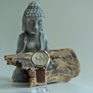 May include: A silver and gold wristwatch with a brown leather band sits on a piece of driftwood in front of a gray concrete statue of a meditating Buddha.
