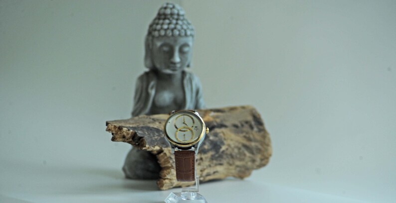May include: A silver and gold wristwatch with a brown leather band sits on a piece of driftwood in front of a gray concrete statue of a meditating Buddha.