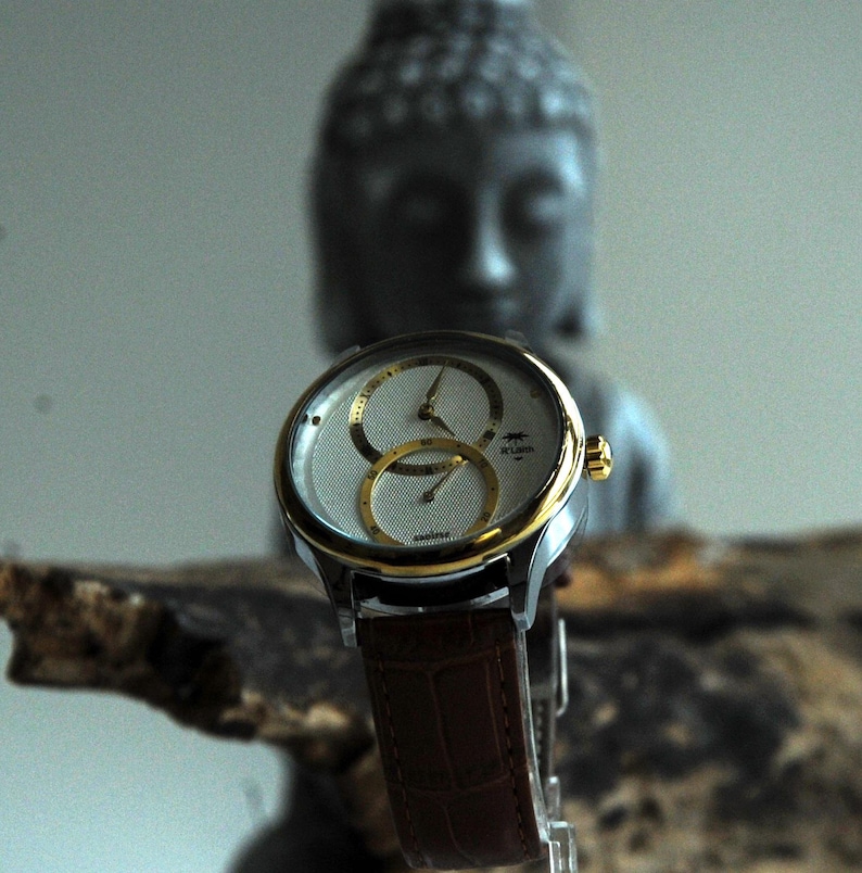 May include: A silver and gold wristwatch with a white face and three subdials. The watch has a brown leather band and is in focus against a blurred background.