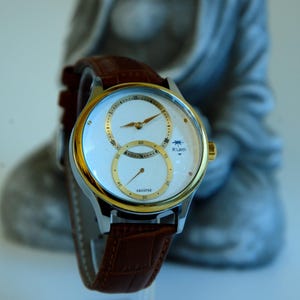 Puede incluir: Un reloj de pulsera analógico blanco con bisel dorado y correa de cuero marrón. La esfera del reloj tiene dos subesferas y el texto "R-Luen" impreso en la esfera.