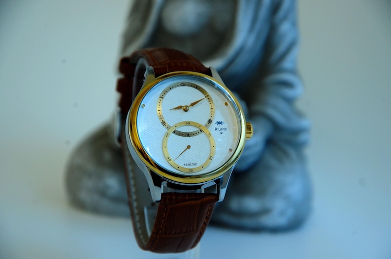 May include: A white analog wristwatch with a gold-tone bezel and brown leather band. The watch face has two sub-dials and the text "R-Luen" printed on the dial.