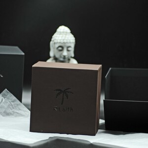 May include: Two black gift boxes with a palm tree logo that says "R'Laith". The boxes are on a white tissue paper surface. A white statue of Buddha is in the background.