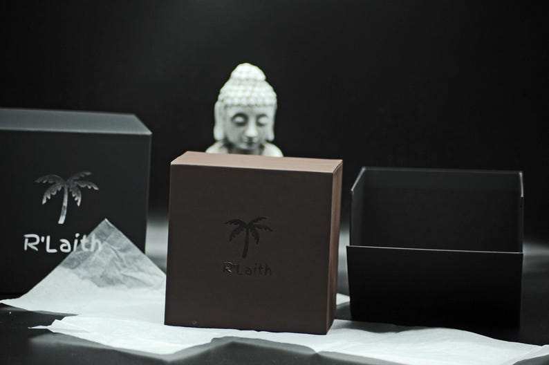 May include: Two black gift boxes with a palm tree logo that says "R'Laith". The boxes are on a white tissue paper surface. A white statue of Buddha is in the background.