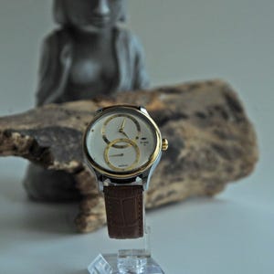 May include: A silver and gold wristwatch with a white face and brown leather band. The watch has two sub-dials and a date window.