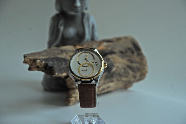May include: A silver and gold wristwatch with a white face and brown leather band. The watch has two sub-dials and a date window.