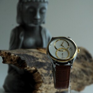 May include: A silver and gold watch with a white face and two round subdials. The watch has a brown leather band and is sitting on a piece of driftwood.