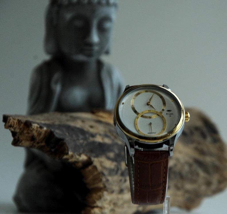 May include: A silver and gold watch with a white face and two round subdials. The watch has a brown leather band and is sitting on a piece of driftwood.