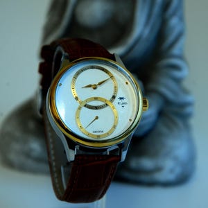 May include: A gold and silver wristwatch with a brown leather band. The watch face has two circular dials with white hands and black Roman numeral markings. The watch face also has the text "sautine" and "R.Luren".