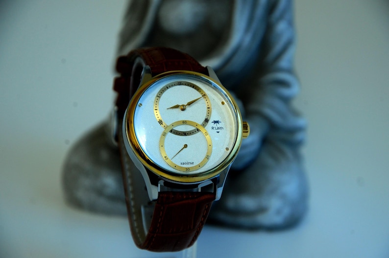 May include: A gold and silver wristwatch with a brown leather band. The watch face has two circular dials with white hands and black Roman numeral markings. The watch face also has the text "sautine" and "R.Luren".