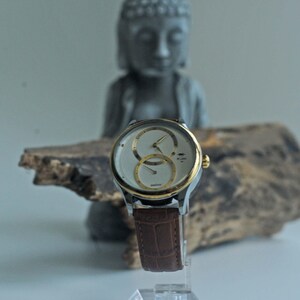 May include: A silver and gold wristwatch with a white face and two subdials. The watch has a brown leather band.