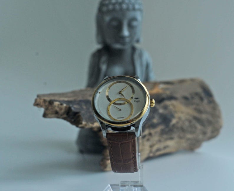 May include: A silver and gold wristwatch with a white face and two subdials. The watch has a brown leather band.