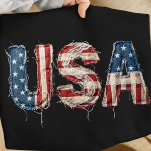 May include: Black textile with the word "USA" in a distressed, patriotic design. The letters are crafted with denim, red and white stripes, and lace, evoking the American flag. The letters are stitched onto the black background.