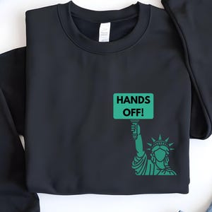 May include: Black sweatshirt with a green graphic of the Statue of Liberty holding a sign that says "Hands Off!"