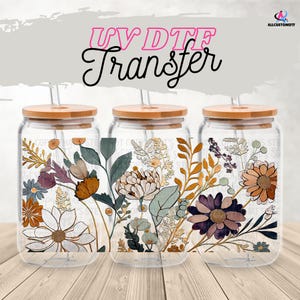 Op de afbeelding: Drie glazen bekers met bloemmotieven, elk met een bamboe deksel en rietje. De bloemmotieven bevatten kleuren als oranje, paars en groen. De tekst "UV DTF Transfer" staat boven de bekers.