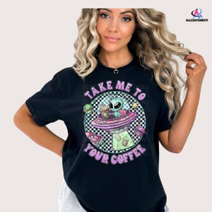 May include: Black t-shirt with a colorful graphic featuring an alien in a spaceship, a coffee cup, and the text "TAKE ME TO YOUR COFFEE". The design includes a checkered background and pastel colors.