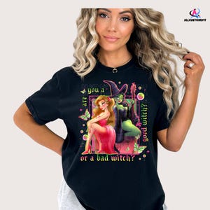 May include: Black t-shirt featuring a colorful graphic of two witches, one in a pink dress and the other in green, with the text "are you a good witch? or a bad witch?" and "DO NOT COPY".