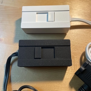 May include: Two rectangular electronic devices, one white and one black, with a recessed rectangular button on the front. Both have attached cables. The white device has a gray cable, and the black device has a black cable. The devices are on a wooden surface.