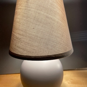 May include: A small, modern table lamp with a white, spherical base and a beige lampshade. The lampshade is conical, with a dark brown trim at the top and bottom. The lamp is designed for indoor use and provides soft, ambient lighting.