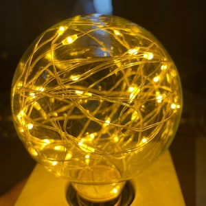 May include: A clear glass orb filled with warm yellow LED string lights. The orb sits on a light-colored, rectangular base. The lights are clustered inside the glass, creating a soft, glowing effect. The overall theme is decorative lighting.