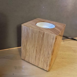 May include: A wooden candle holder with a square shape and a light brown finish. The holder has a circular white insert for a candle. The wood grain is visible on the surface.