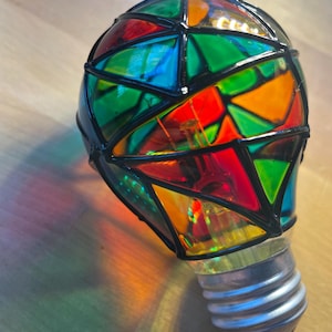 May include: A light bulb with a stained glass design. The glass is in shades of red, orange, green, and blue. The bulb is black outlined and has a silver screw-in base.