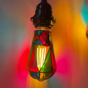 May include: A stained glass light bulb with a black socket and a warm white filament. The bulb is decorated with red, green, blue, and yellow glass pieces.