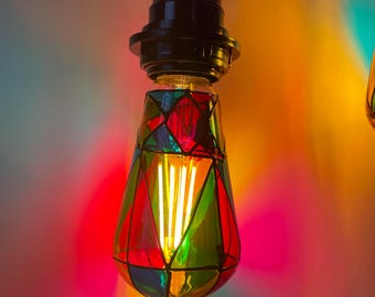 May include: A stained glass light bulb with a black socket and a warm white filament. The bulb is decorated with red, green, blue, and yellow glass pieces.