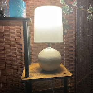 May include: A small table lamp with a white lampshade and a spherical, textured base. The lamp sits on a small wooden table with black legs. A blue glass candle holder is visible on a shelf to the left.