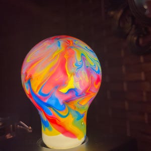 May include: A colorful light bulb with a swirling pattern of red, yellow, blue, and green paint. The bulb has a white base and is set against a dark background. The bulb is a decorative item.