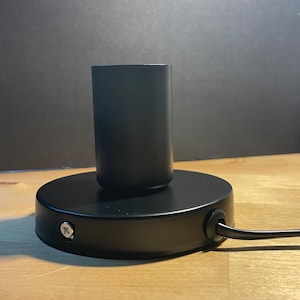 May include: A black lamp base with a cylindrical socket and a round base. A black power cord extends from the base. The lamp base is on a wooden surface.