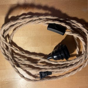 May include: A coiled, beige rope light cord with a black inline switch, a black bulb socket, and a black plug. The cord is made of twisted natural fibers, suggesting a rustic or industrial aesthetic. The components are black.