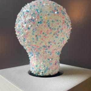 May include: A decorative light bulb covered in white, pink, and blue glitter. The bulb is sitting on a white surface with a black base. The bulb has a round top and a bulbous shape.