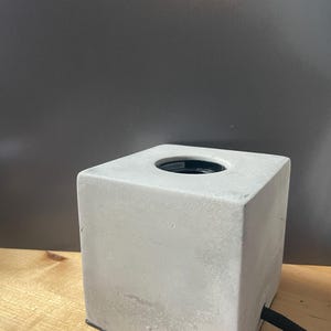 May include: A square, light gray concrete tissue box with a circular opening on top. A black cord extends from the side. The tissue box sits on a wooden surface against a dark gray background.