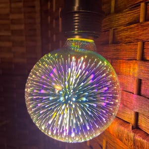 May include: A decorative, round light bulb with a black base. The bulb is clear with a colorful, starburst-like design in shades of purple, yellow, and white. The bulb is hanging in front of a woven, brown background.