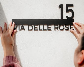 Puede incluir: Un letrero de número de casa de metal negro con el número 15 y el texto "VIA DELLE ROSE" en una pared blanca.