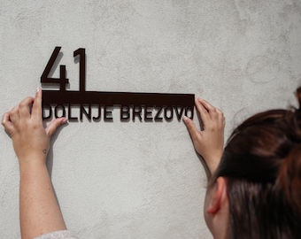 Puede incluir: Una persona está instalando un letrero de número de casa negro con el número 41 y el texto "DOLNJE BREZOVO" en una pared blanca.