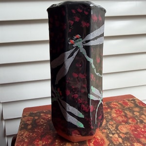May include: A tall, black ceramic vase with a rectangular shape. The vase features a glossy finish and is decorated with white and gray dragonfly illustrations and red floral accents. The base of the vase is a reddish-brown color.