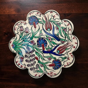 May include: A scalloped ceramic plate with a white base featuring a hand-painted floral pattern. The design includes blue, red, and green tulips and leafy stems with raised paint textures. The plate has a distinct wavy edge and a glossy finish.