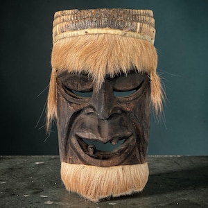 May include: A carved wooden mask with a dark brown face, closed eyes, and a wide, toothy grin. The mask features a band of light-colored fur around the forehead and chin, with longer fur framing the face. The mask is likely of cultural or tribal origin.