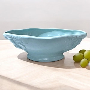 May include: A light blue ceramic bowl with a decorative grape design. The bowl has a wide, oval shape and a raised base.
