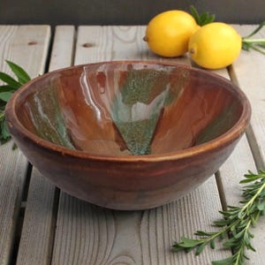 May include: A handmade ceramic bowl with a glossy finish. The bowl features a blend of brown and green hues, creating a unique, organic appearance. The bowl is round and has a wide opening, suitable for serving or display. Two yellow lemons and sprigs of rosemary are in the background.