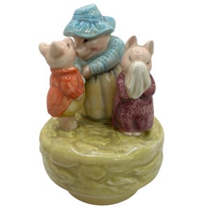 May include: A ceramic figurine featuring three pig characters. One pig wears a red coat, another a purple dress, and the third a blue hat and shirt. The base is a light green color, and the figurine has a glossy finish.