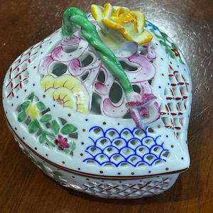 May include: A white ceramic basket-shaped trinket box with a pierced design. The lid features a green handle and a yellow rose. The box is decorated with pink, yellow, and blue floral accents and a brown trim.