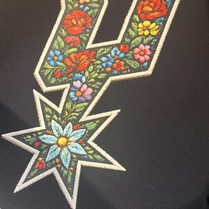 May include: Black t-shirt featuring a floral design in the shape of a stylized letter "U" and a star. The design incorporates colorful flowers in red, orange, yellow, blue, and pink, with a white outline.