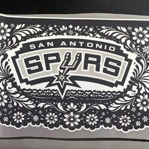 May include: A black and white sticker with the text "San Antonio Spurs". The team name is set against a gray and black background, surrounded by a floral design. A star and spur graphic are also included.