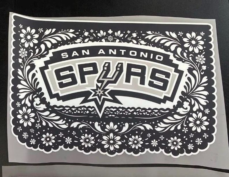 May include: A black and white sticker with the text "San Antonio Spurs". The team name is set against a gray and black background, surrounded by a floral design. A star and spur graphic are also included.