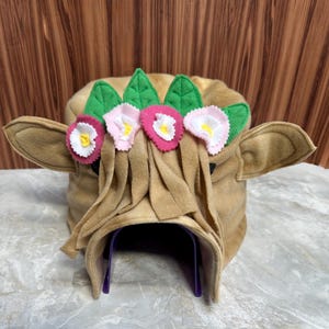 May include: A brown felt pet bed shaped like a cow's head with pink and white felt flowers and green leaves around the head. The bed has a purple tunnel entrance.