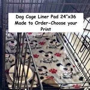 May include: A white dog cage liner pad measuring 61 cm by 91 cm, featuring a repeating pattern of cartoon dogs and red paw prints with the text I Woof You, placed inside a black wire metal crate with a white bowl.